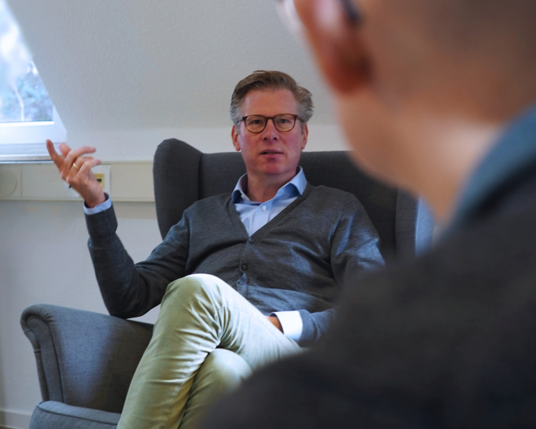 Ein Mann sitzt in einem Sessel mit überschlagenen Beinen und befindet sich in einem Gespräch.