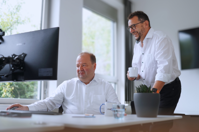 Zwei Männer arbeiten gemeinsam an einem Computer in einem hellen Büro; einer sitzt am Bildschirm, der andere steht daneben mit einer Tasse in der Hand.